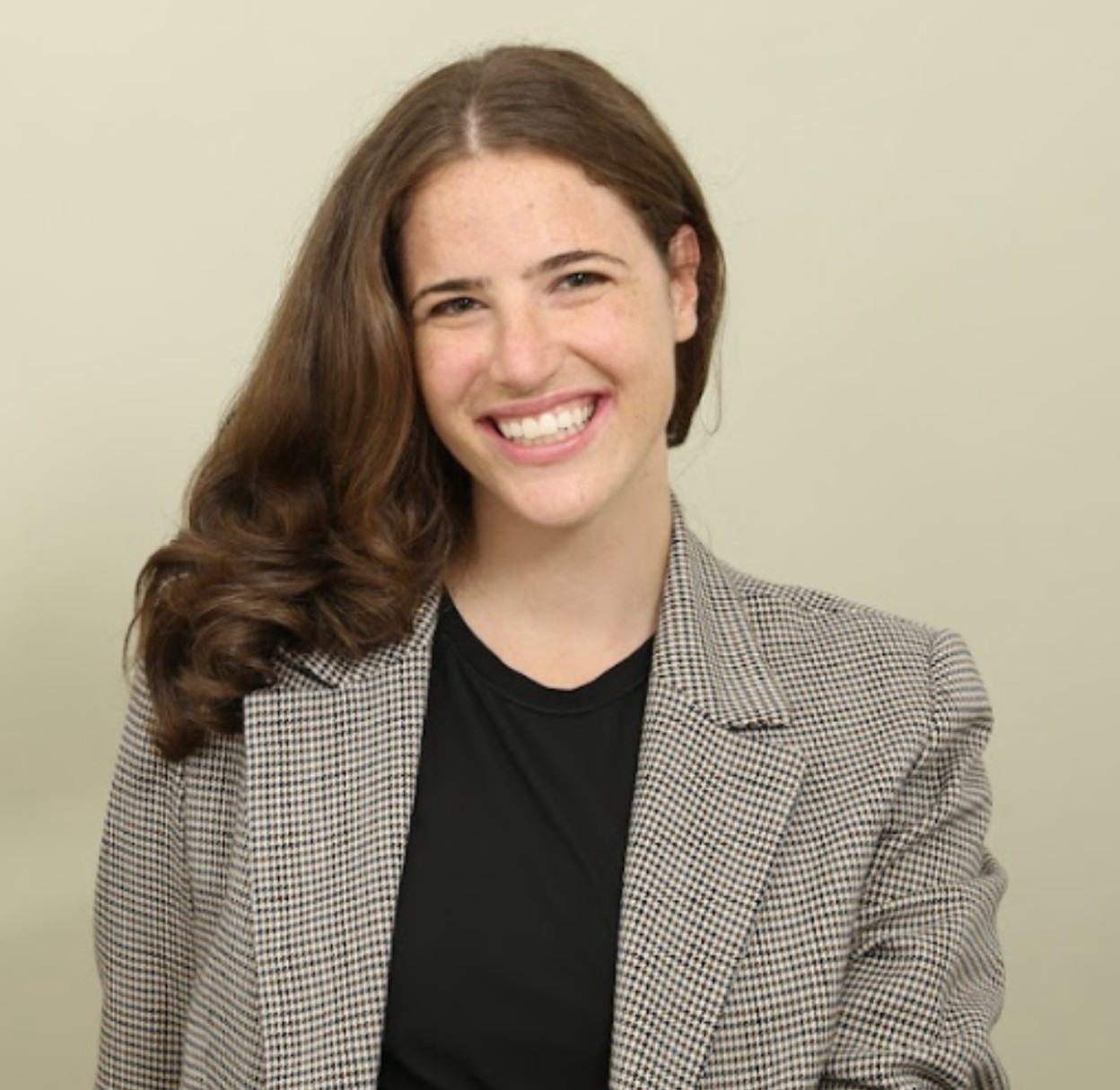 Professional headshot of a smiling woman with long brown hair wearing a black top and checkered blazer.