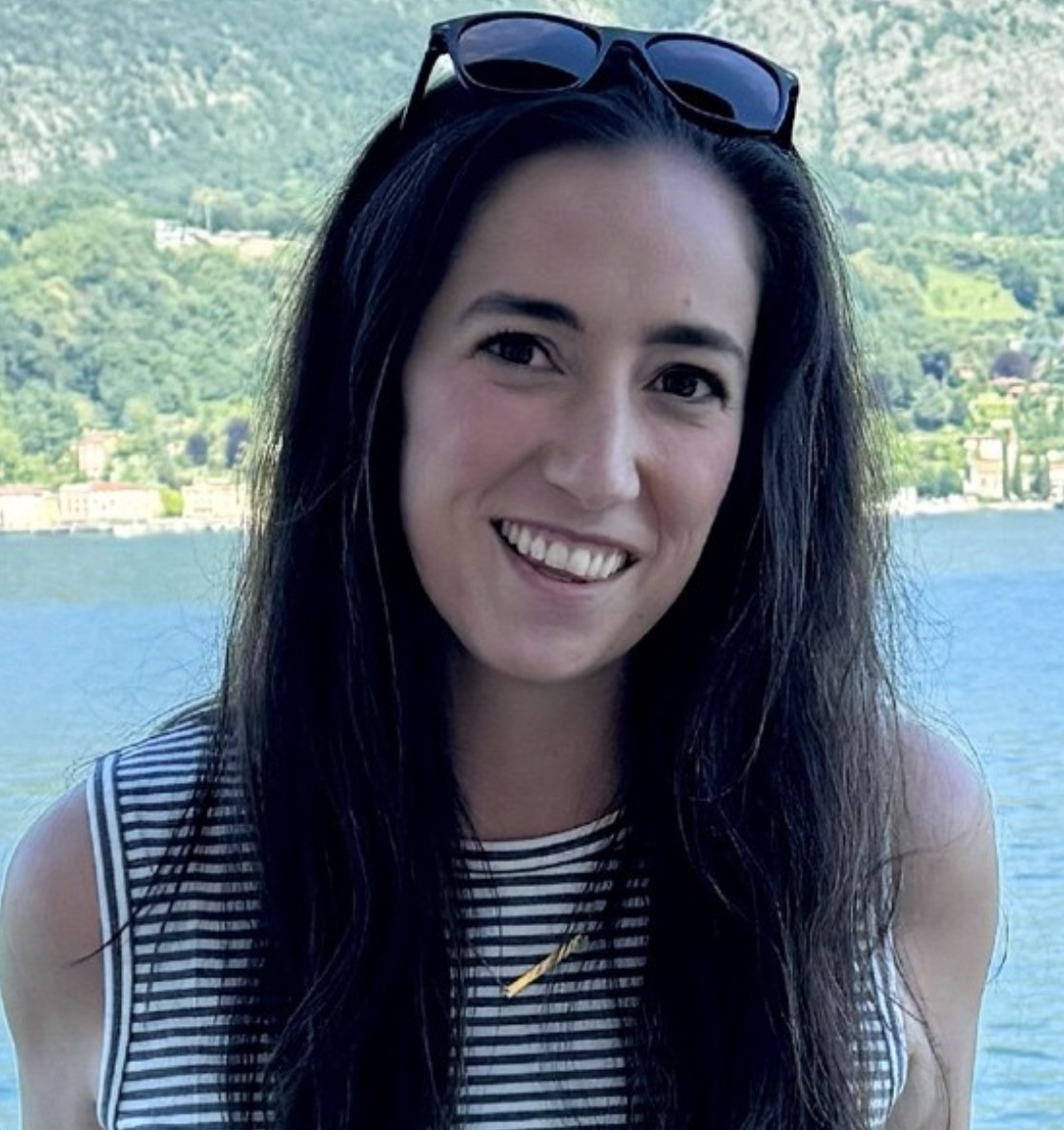 Portrait of a smiling woman with long dark hair standing outdoors near a lake with green mountains in the background. She is wearing a black and white striped sleeveless top and sunglasses resting on her head.