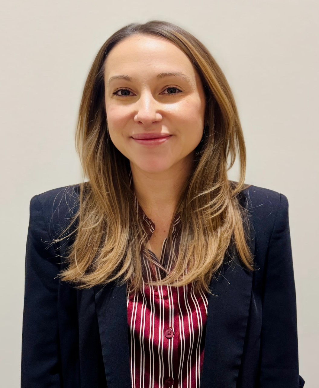 Professional headshot of a woman with long light brown hair wearing a black blazer and red striped blouse.