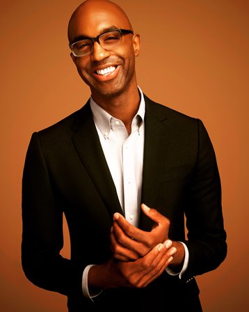 joseph nelson in a white shirt and black blazer, smiling with an burnt orange background