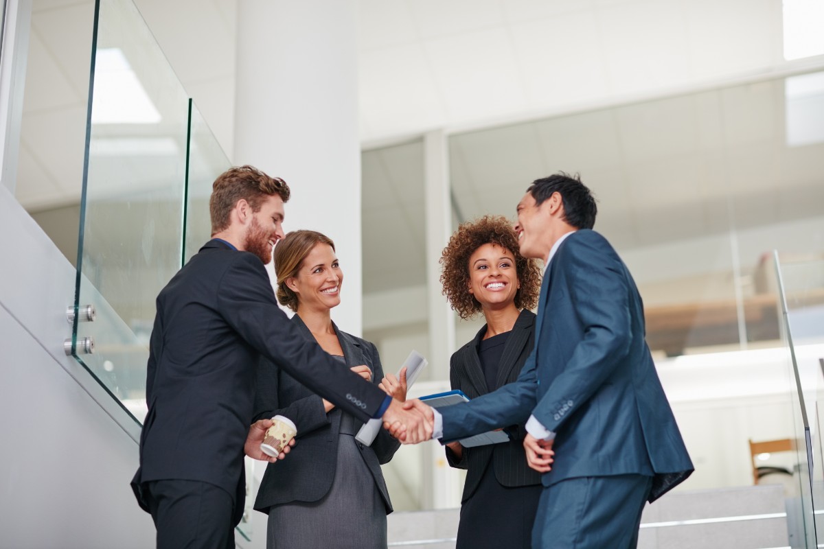 A group of working professionals in an office setting standing together smiling while holding a laptop and tablet