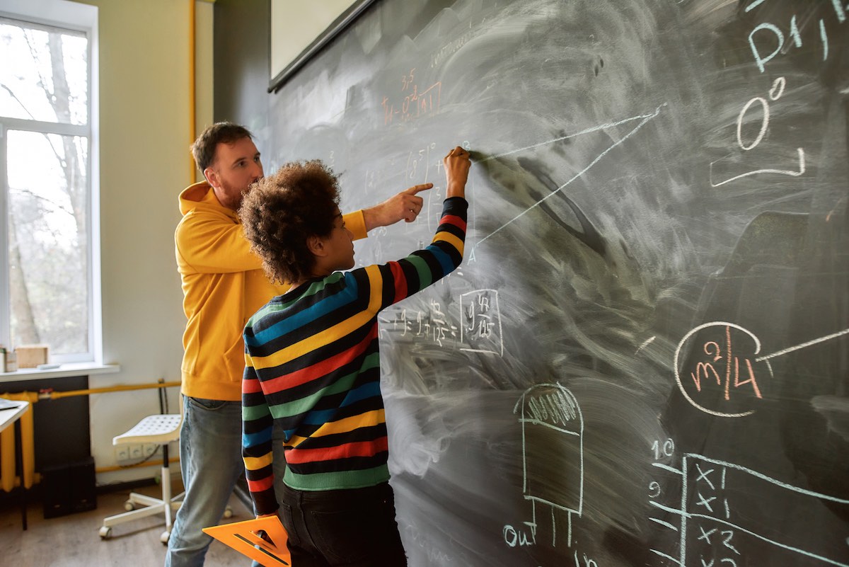 Young african american child with an afro and a colorful sweater standing at a chalkboard with a white teacher. Both subects are working on mathematical equations on the chalkboard