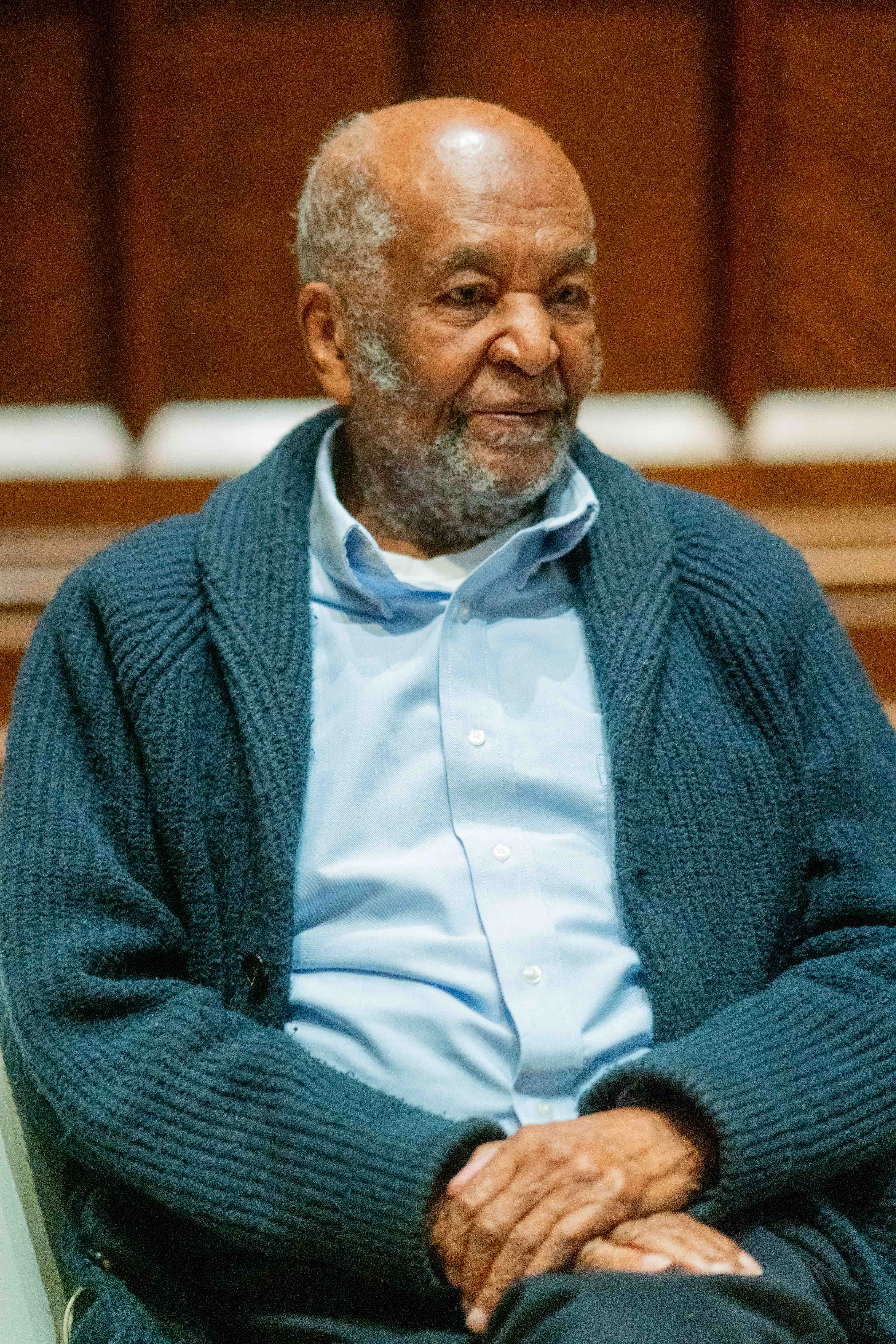 An elderly black man in a blue shirt and teal cardigan is sitting down with his hands folded on his lap