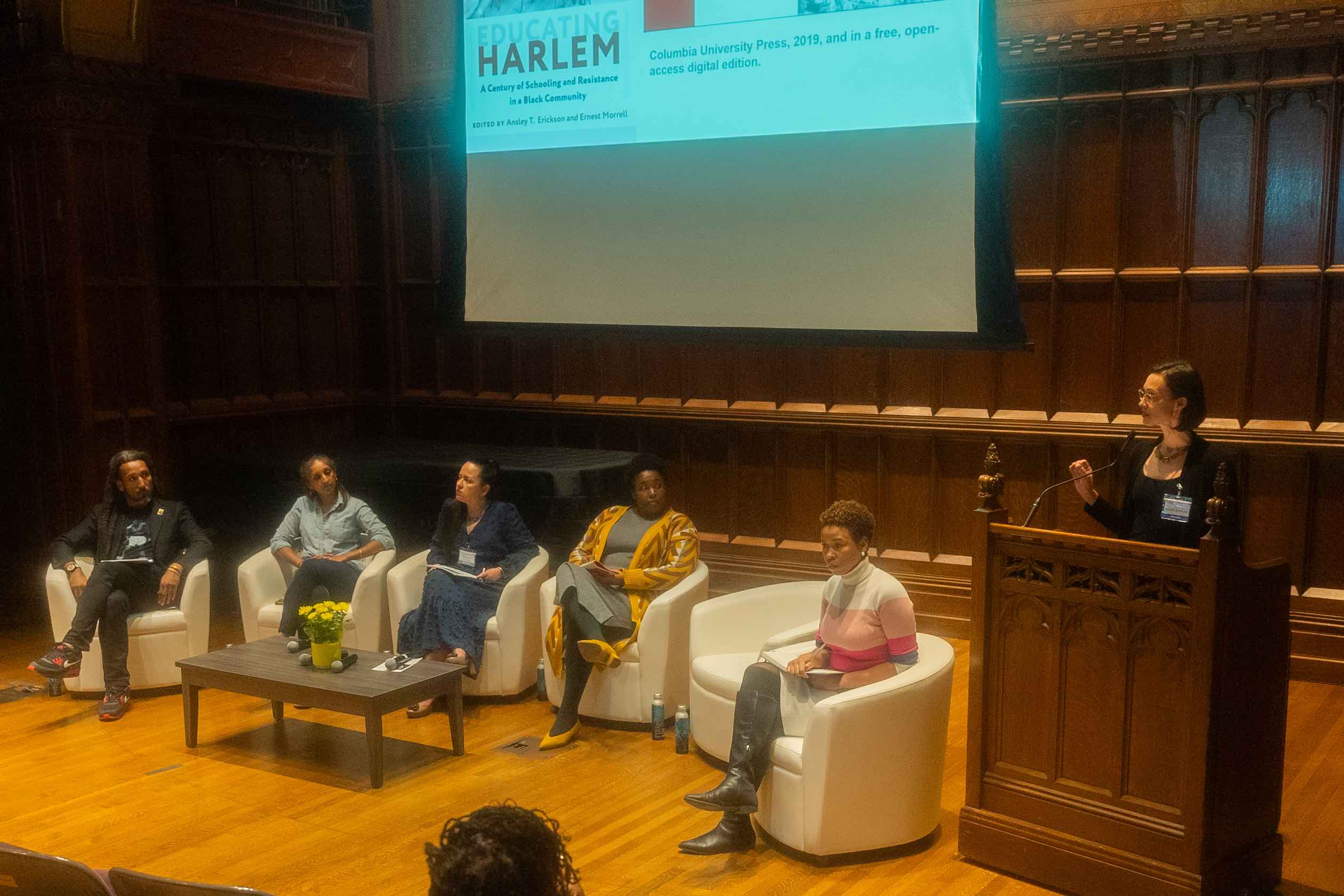 Five people on a stage sit on white chairs, looking at a sixth person standing behind a podium