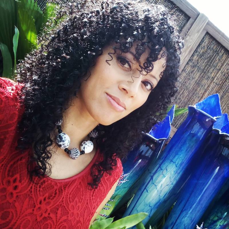 Jacqueline cofield smiling softly at the camera. She is in a yard. She has light skin, long dark curly hair and is wearing a red shirt with a statement necklace