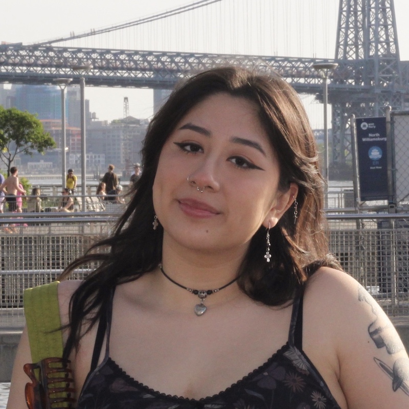 Sofia Rosario looking at the camera with a large bridge in the background. She has wavy dark hair and bold winged eyeliner