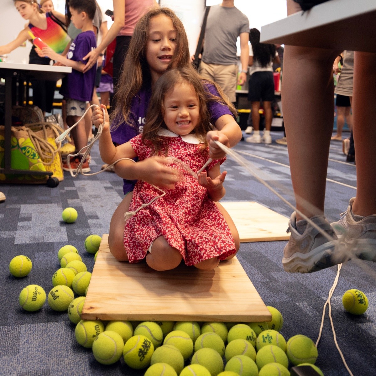 Two girls play on a homemade sled, constructed with a plank, reigns and tennis balls.