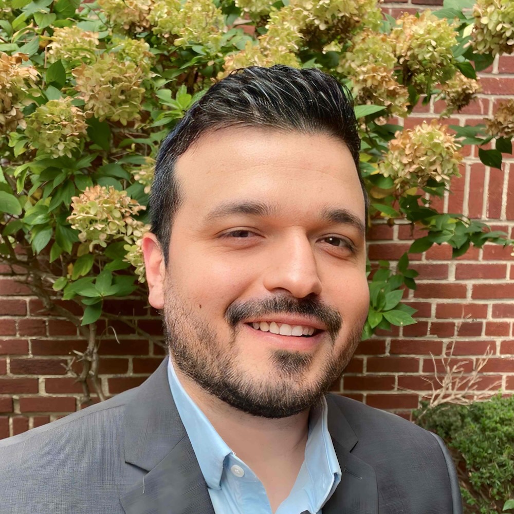 Eddie Rivero, a man with a beard, is smiling in front of a brick wall with greenery