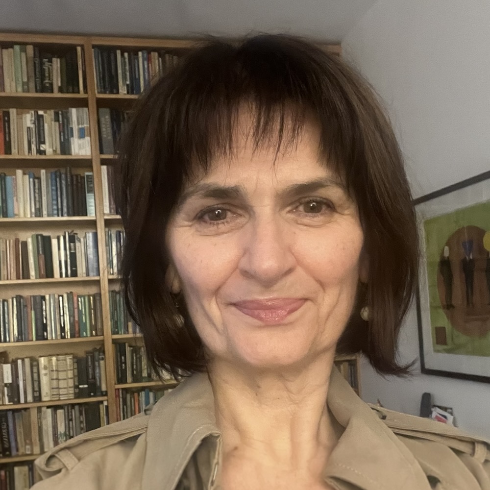 eleni nastioupoulos, a woman with short brown hair and bangs, is smiling infront of a bookshelf