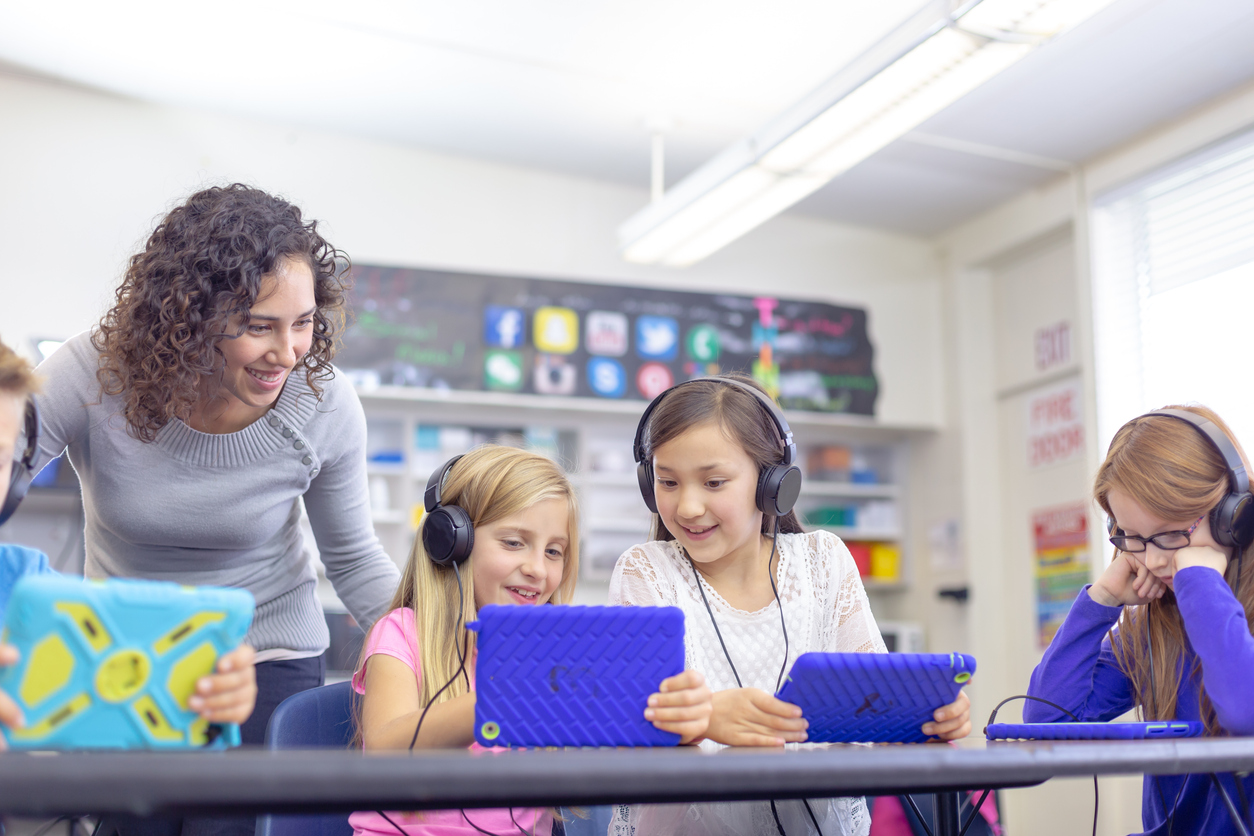A group of elementary age students sit at their school desks and smile while using tablet computers with headphones to complete an assignment in class. A young female teacher is checking the school children's progress and providing assistance.