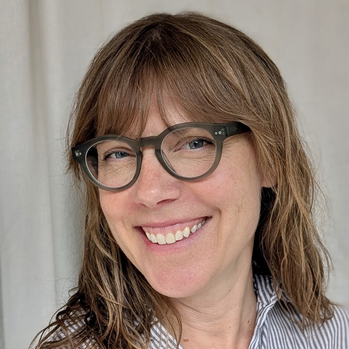 A woman with fair skin, brown hair with bangs, and glasses smiles at the camera