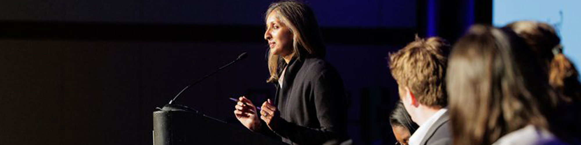 close up of woman at a podium with a microphone with a few people next to her and looking at her