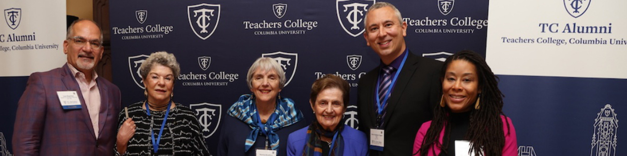 group of six people smiling and wearing name tags, standing in a row in front of a teachers college sign