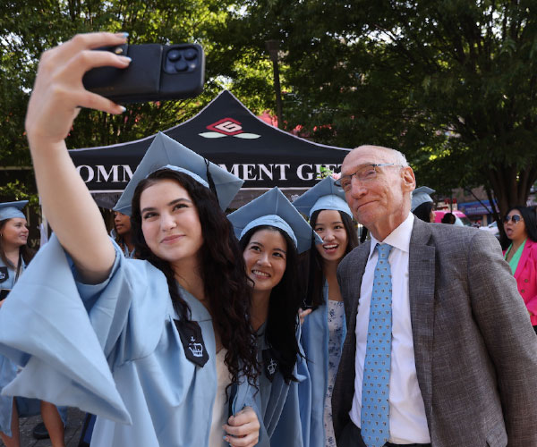 three students wearing graduation cap and gown and president Thomas Bailey taking a selfie in an outdoor setting