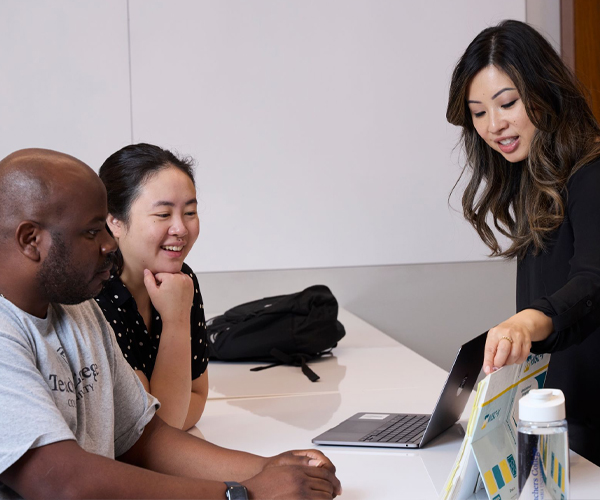 two people seated and one person standing all looking at a laptop on the table