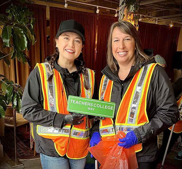 two people standing and smiling, wearing reflective work vests and plastic gloves, holding a trashbag