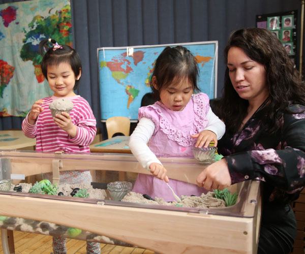 two children and an adult standing with their hands reaching into a sandbox with gardening objects in it