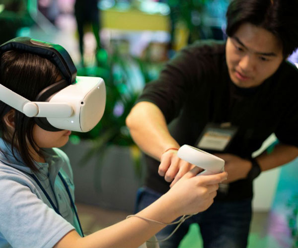 person wearing virtual reality goggles holding a handheld device out in front of them with person next to them touching the handheld device