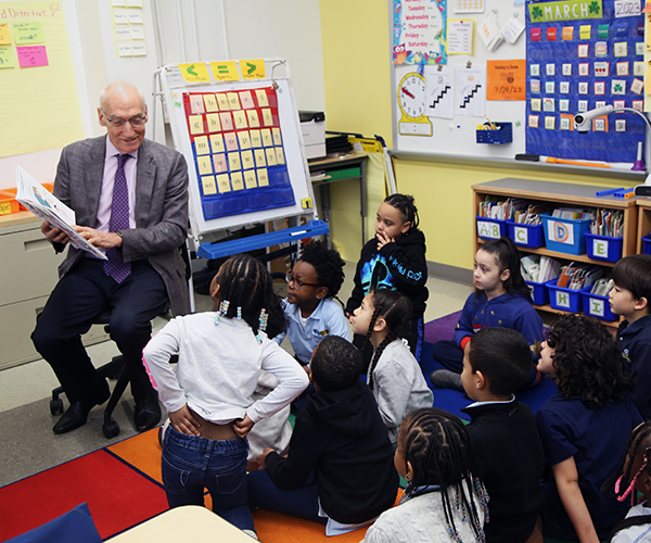 president Tom Bailey seated and holding a book with a group of young students seated on the floor