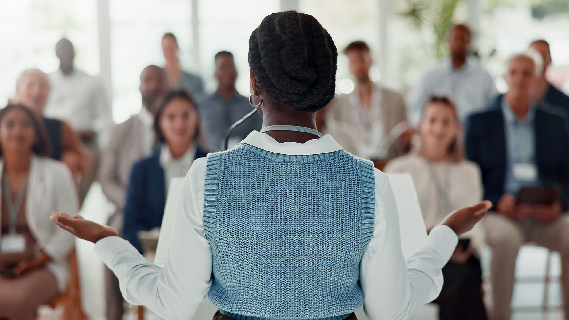 View of the back of a woman standing and speaking in front of a large group