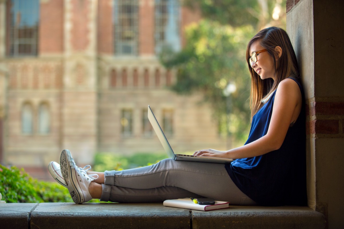 A woman sitting on a stone ledge outdoors while working on a laptop