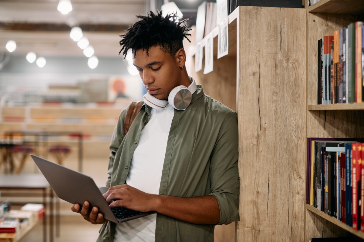 A man with headphones around his neck leaning against a bookshelf holding an open laptop